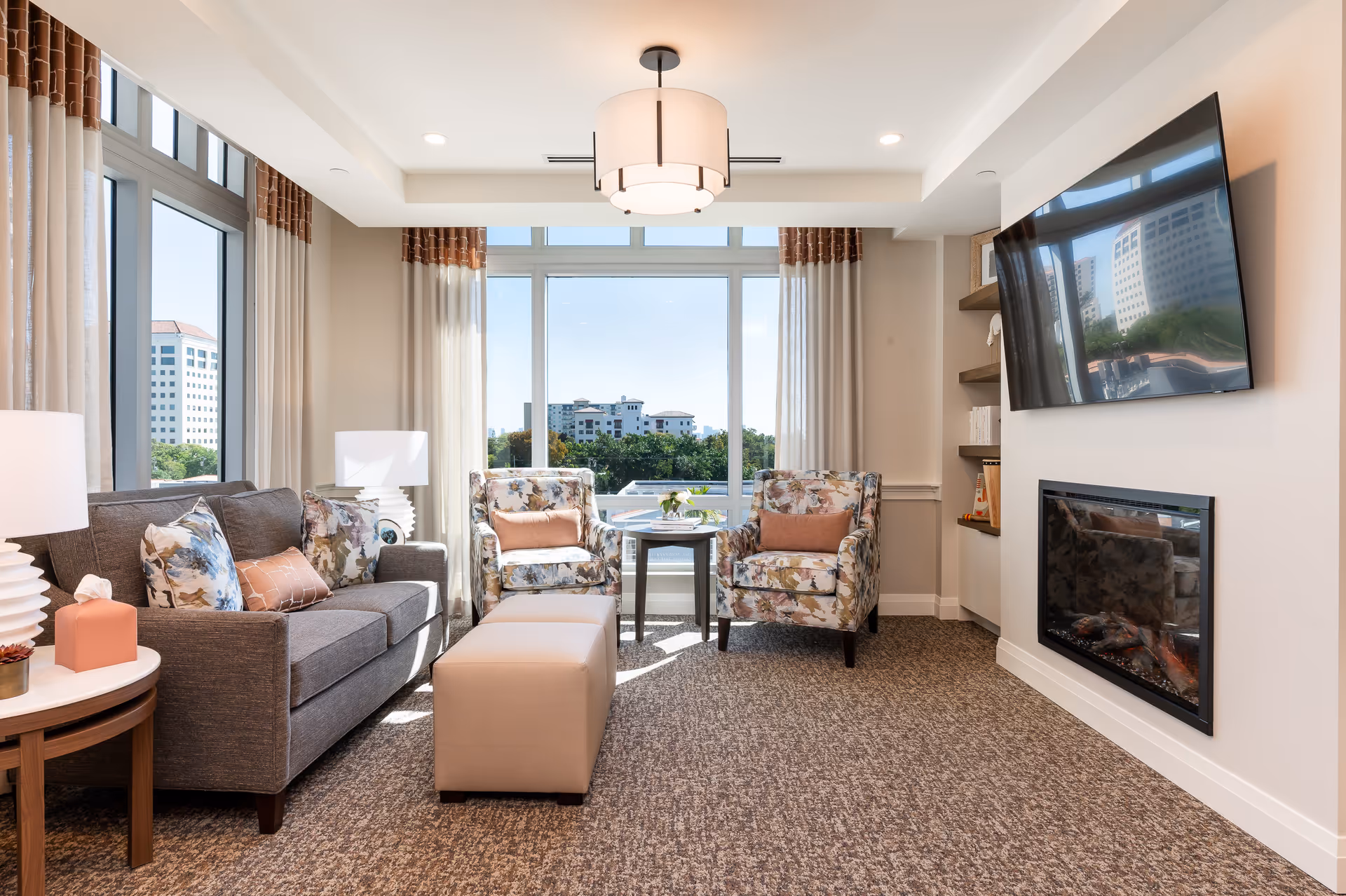 A bright and cozy living room with a large window showing a cityscape view. The room features a gray sofa with patterned and solid pillows, two floral armchairs with pink cushions, a beige ottoman, a round side table with a lamp and tissue box, a wall-mounted flat-screen TV above a modern electric fireplace, and built-in shelves with decorative items and books. The room is carpeted and has beige walls with white trim and ceiling lights.