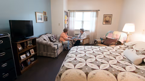 A cozy room with a large bed covered in a patterned quilt, a sofa, an armchair with a lamp beside it, and a TV on a wooden stand. Two elderly women are sitting near the window, engaged in conversation.