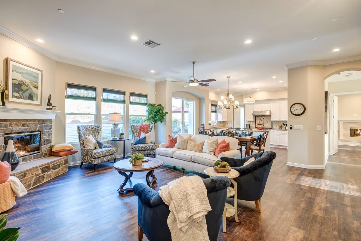 A bright and spacious living room area with a stone fireplace on the left, two patterned armchairs, a beige sofa with multiple cushions, and two dark blue armchairs with a white knitted throw. A round wooden coffee table and a small side table with plants are in the center. The room has large windows with blinds, wooden flooring, and recessed ceiling lights. In the background, there is a dining area with a wooden table and chairs, a chandelier, and a kitchen with white cabinets and stainless steel appliances.