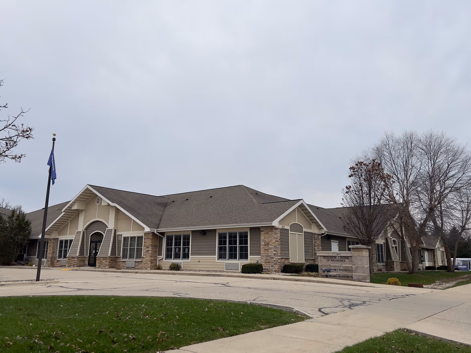 Mission Creek Senior Living single-story building with stone accents, a flagpole, and a driveway under an overcast sky.