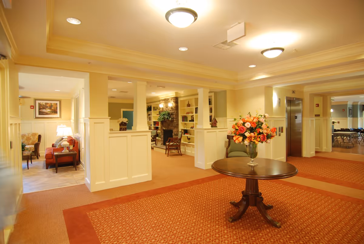 A well-lit interior common area with beige walls and white wainscoting, featuring a round wooden table with a large vase of colorful flowers in the center. The space includes a seating area with chairs and a fireplace surrounded by built-in shelves filled with decorative items. An elevator is visible on the right side, and there are additional seating and tables in the background.