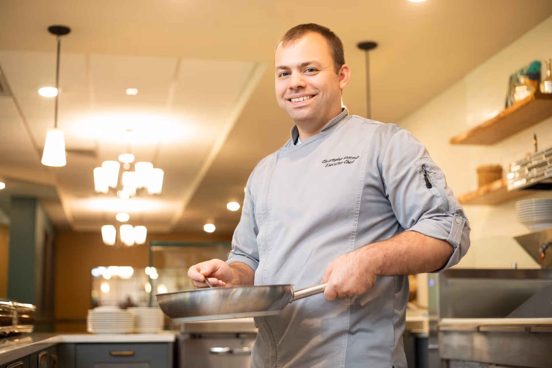 A smiling male chef wearing a light gray chef's jacket stands in a kitchen holding a frying pan. The kitchen has warm lighting with hanging pendant lights and shelves with plates and kitchen items in the background.