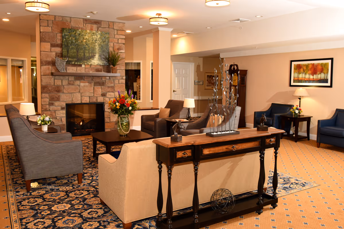 A cozy senior living facility lounge area with a stone fireplace, a painting above it, and a vase of colorful flowers on a coffee table. The room features comfortable armchairs and sofas arranged on a patterned carpet, with side tables holding lamps and small flower arrangements. There is a wooden console table with decorative items and a grandfather clock in the background. The walls are painted beige with framed artwork.
