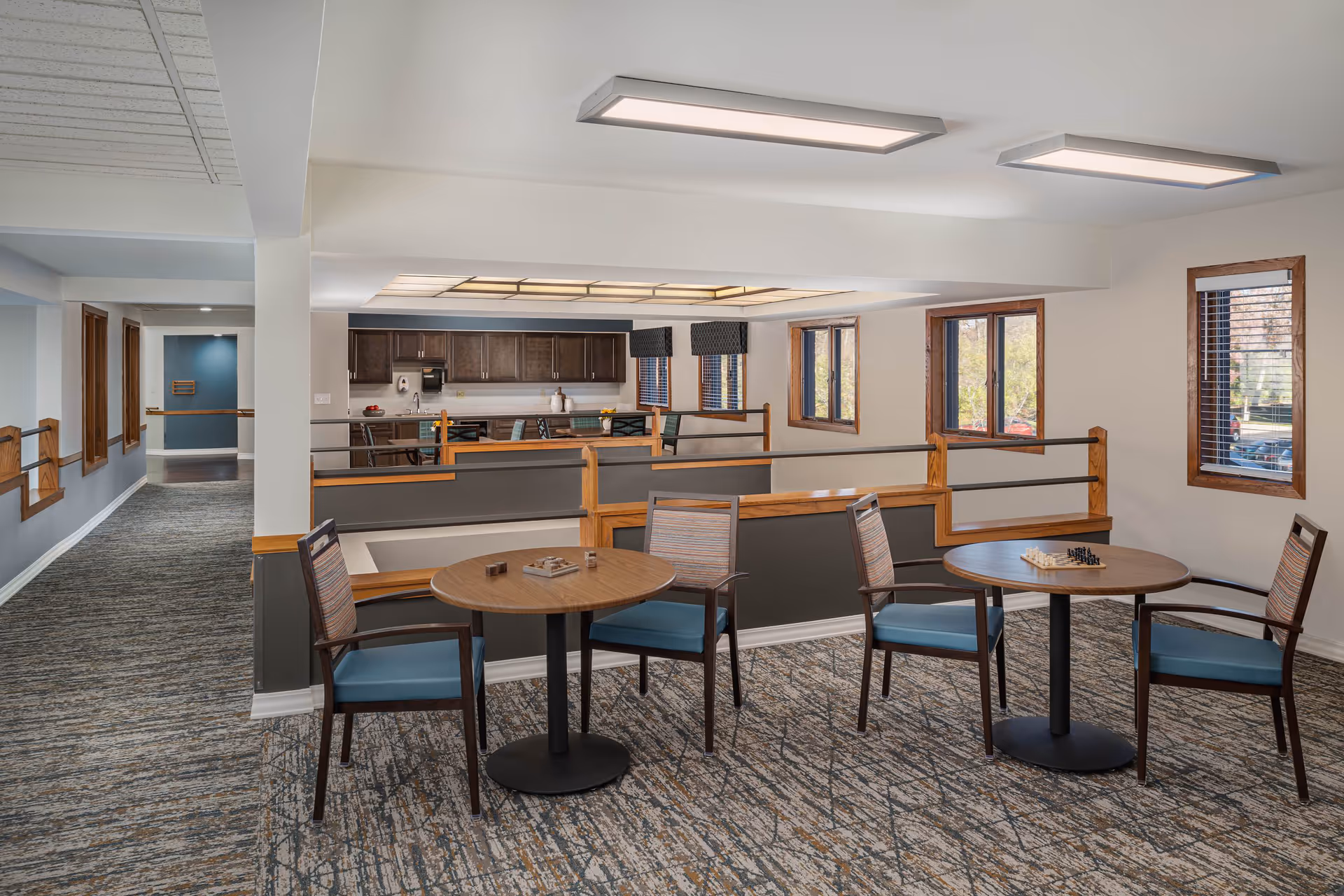 Interior view of a senior living community common area with two round tables, each surrounded by two chairs. The tables have board games on them. The space has carpeted floors, large windows with wooden frames, and a kitchen area with dark cabinets in the background.