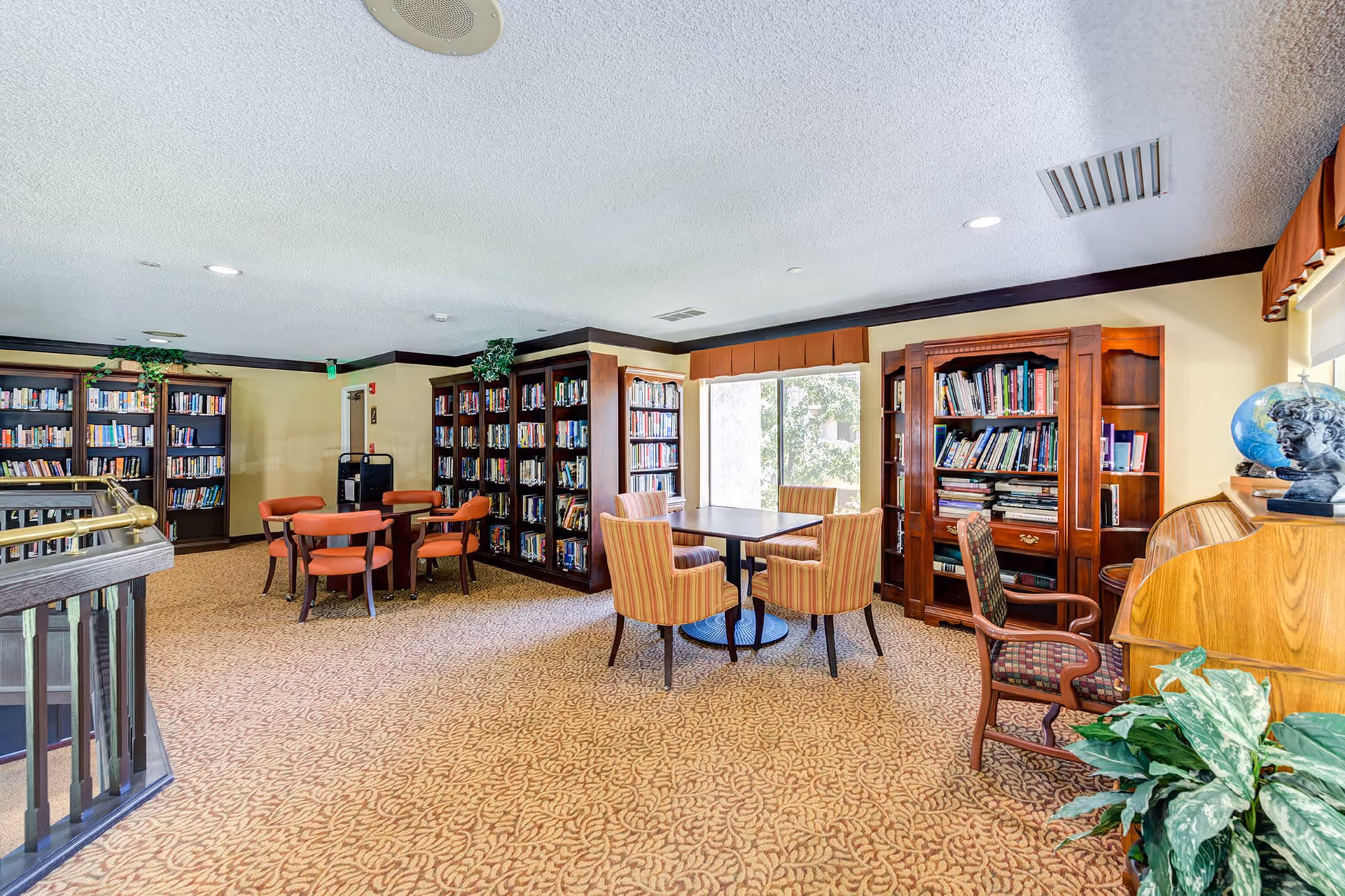 A cozy senior living facility library with multiple wooden bookshelves filled with books, several seating areas including round tables with orange and striped chairs, a large window letting in natural light, and a wooden desk with a globe and bust on top. The room has beige walls, patterned carpet, and some green plants for decoration.
