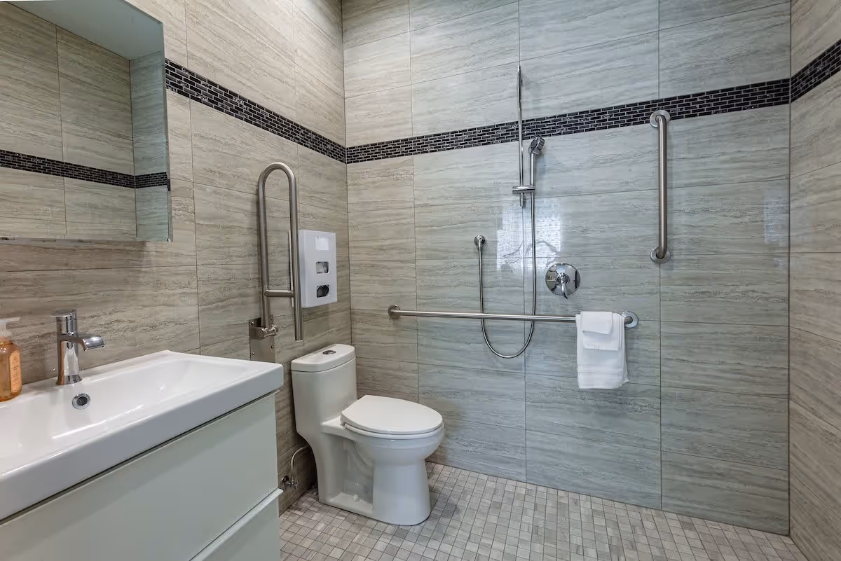 A modern bathroom with beige tiled walls and floor. It features a white sink with a chrome faucet, a toilet, and a walk-in shower area with a handheld showerhead. There are multiple stainless steel grab bars installed around the toilet and shower area for accessibility, and a white towel hangs on one of the bars.