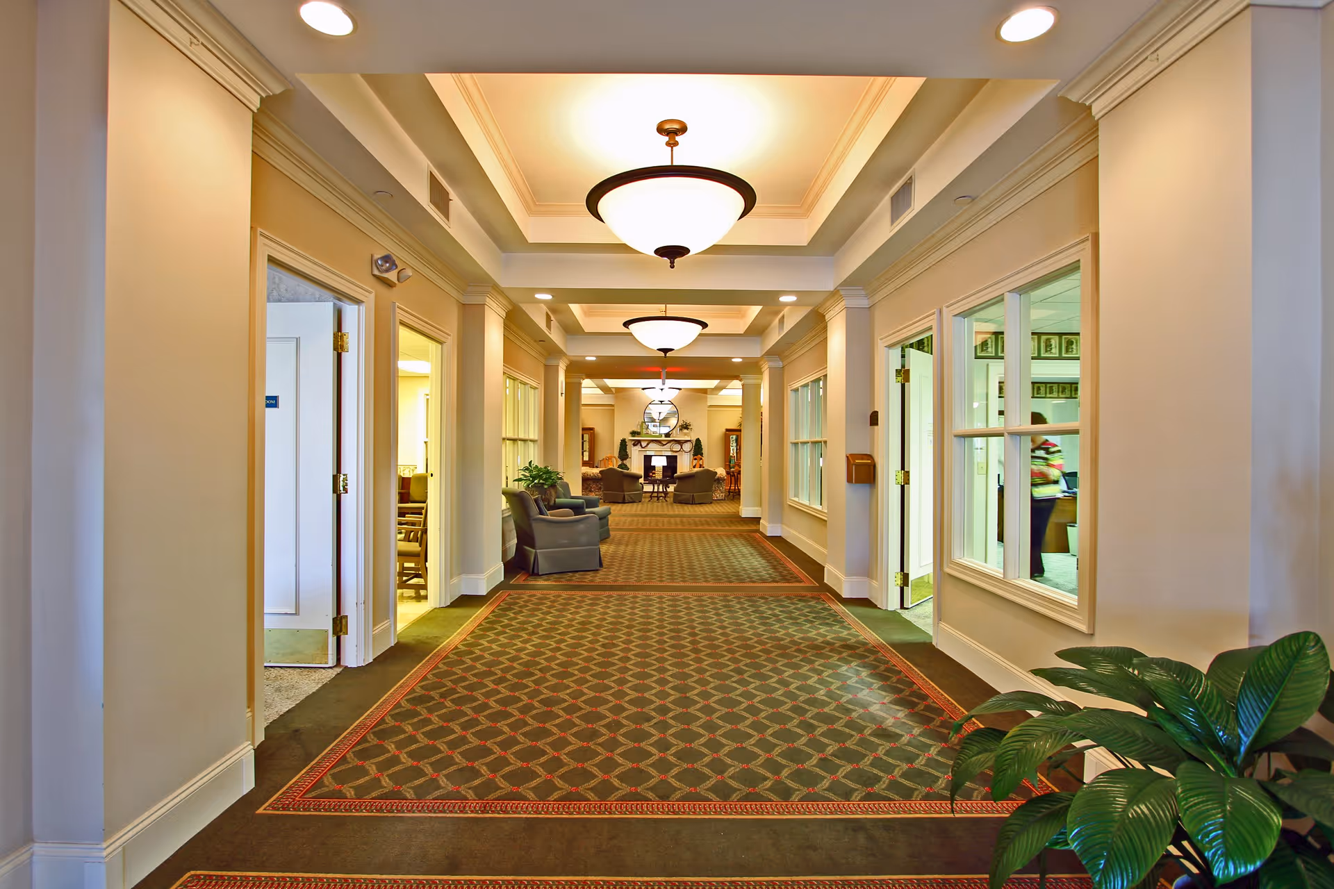 A well-lit hallway in The Garden House of Anderson with patterned carpet, beige walls, and white trim. The hallway features ceiling lights and several doorways on both sides, some open revealing rooms with chairs. At the end of the hallway is a cozy sitting area with armchairs and a fireplace. A green leafy plant is visible in the foreground on the right side.