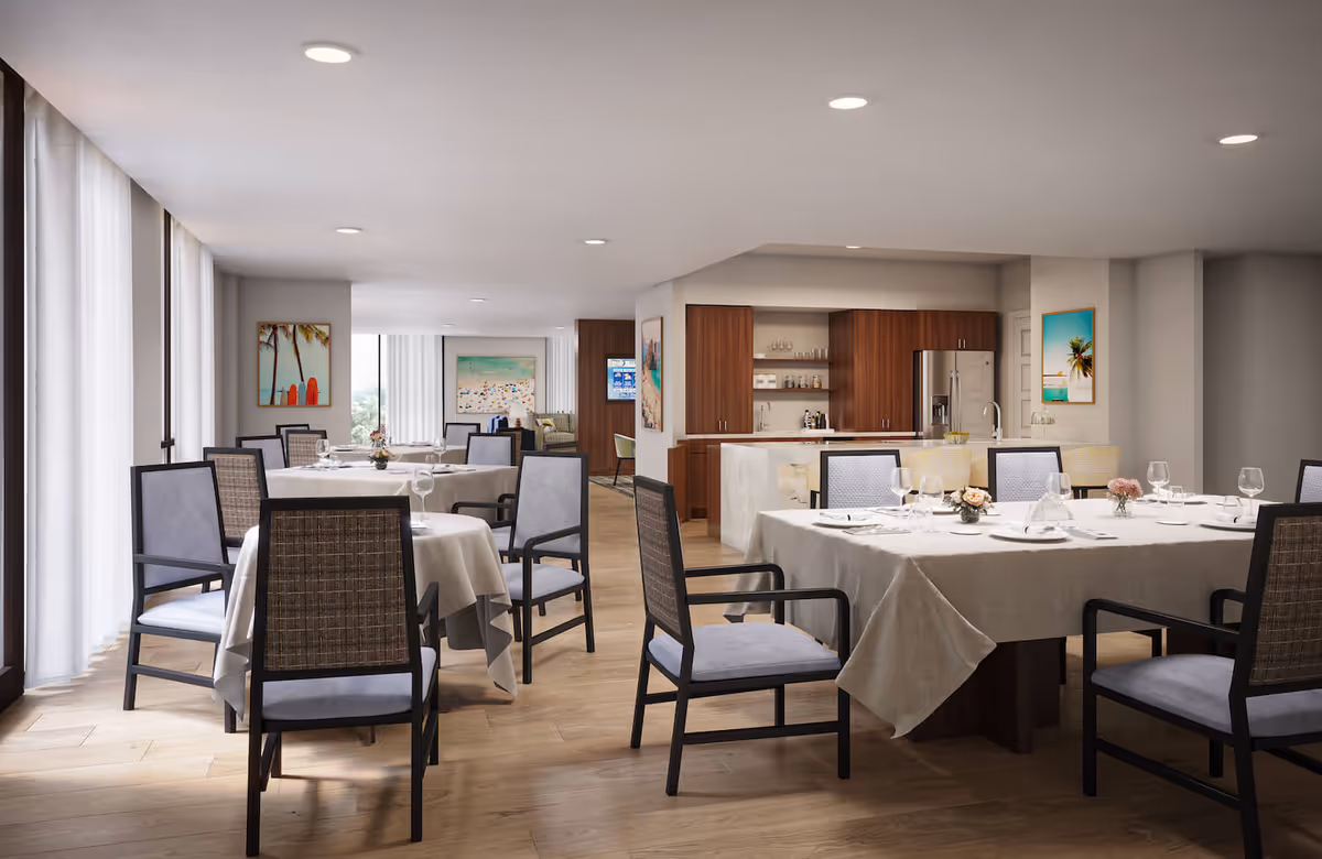 A bright dining room with several round and rectangular tables covered with white tablecloths, set with glassware and small floral centerpieces. The room features wooden flooring, large windows with sheer white curtains, and modern chairs with cushioned seats and woven backs. In the background, there is a kitchen area with wooden cabinets, a refrigerator, and a countertop with bar stools. The walls are decorated with colorful beach-themed paintings.