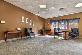 A spacious room with beige walls and a carpeted floor featuring a subtle pattern. The room contains two upholstered armchairs and a small round wooden table near a window with blue valance curtains. On the left side, there is a wooden console table with books and decorative items. The ceiling has recessed lighting and a smoke detector.