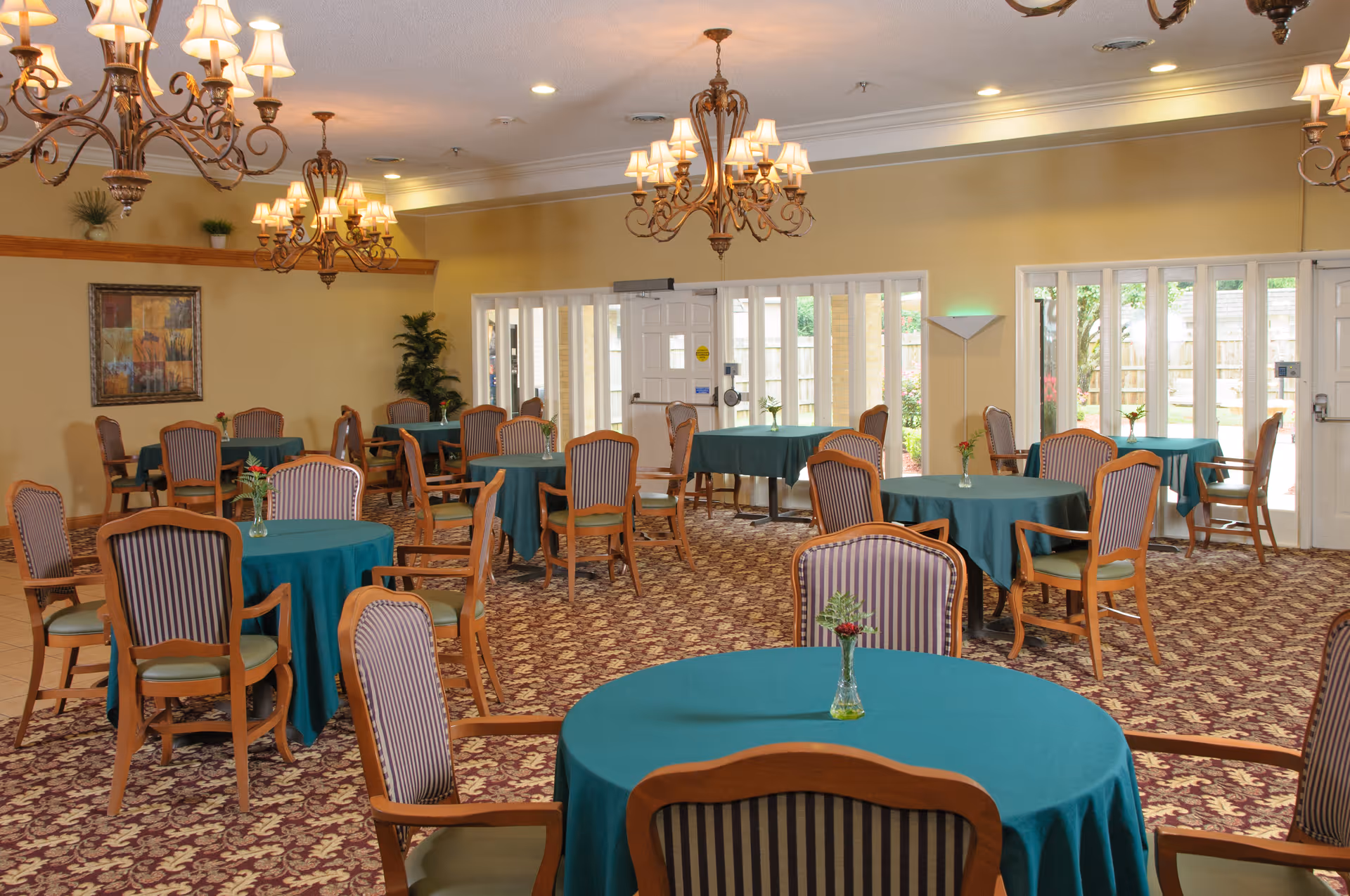 Spacious dining room with round tables covered in teal tablecloths, wooden chairs, chandeliers, and large windows.