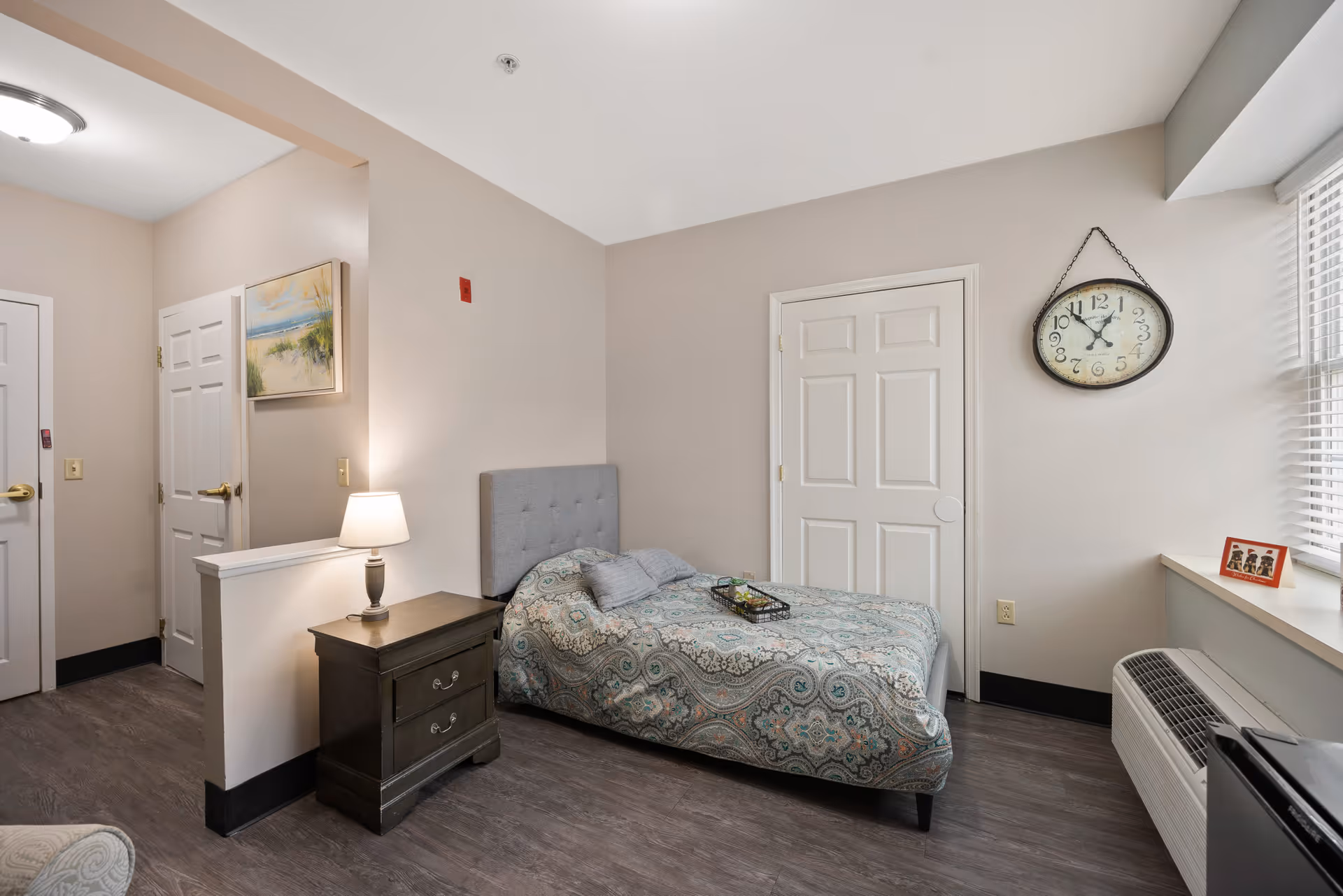 A tidy single-occupancy bedroom with a bed, nightstand and lamp, wall clock, and a window air conditioner.