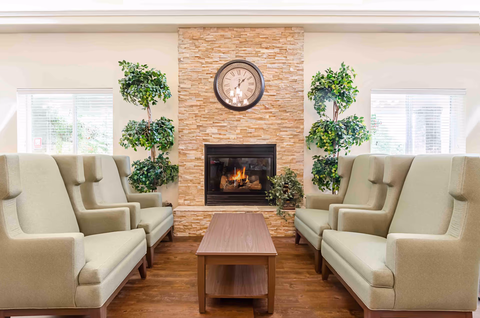 A cozy seating area with two beige upholstered armchairs on each side facing each other, a wooden coffee table in the center, a stone fireplace with a fire burning, a round wall clock above the fireplace, and two tall green potted plants on either side of the fireplace. Two windows with white blinds flank the fireplace.