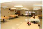 Large multipurpose dining/activity room with several rectangular tables covered in peach tablecloths, chairs, floral centerpieces, and overhead fluorescent lights.