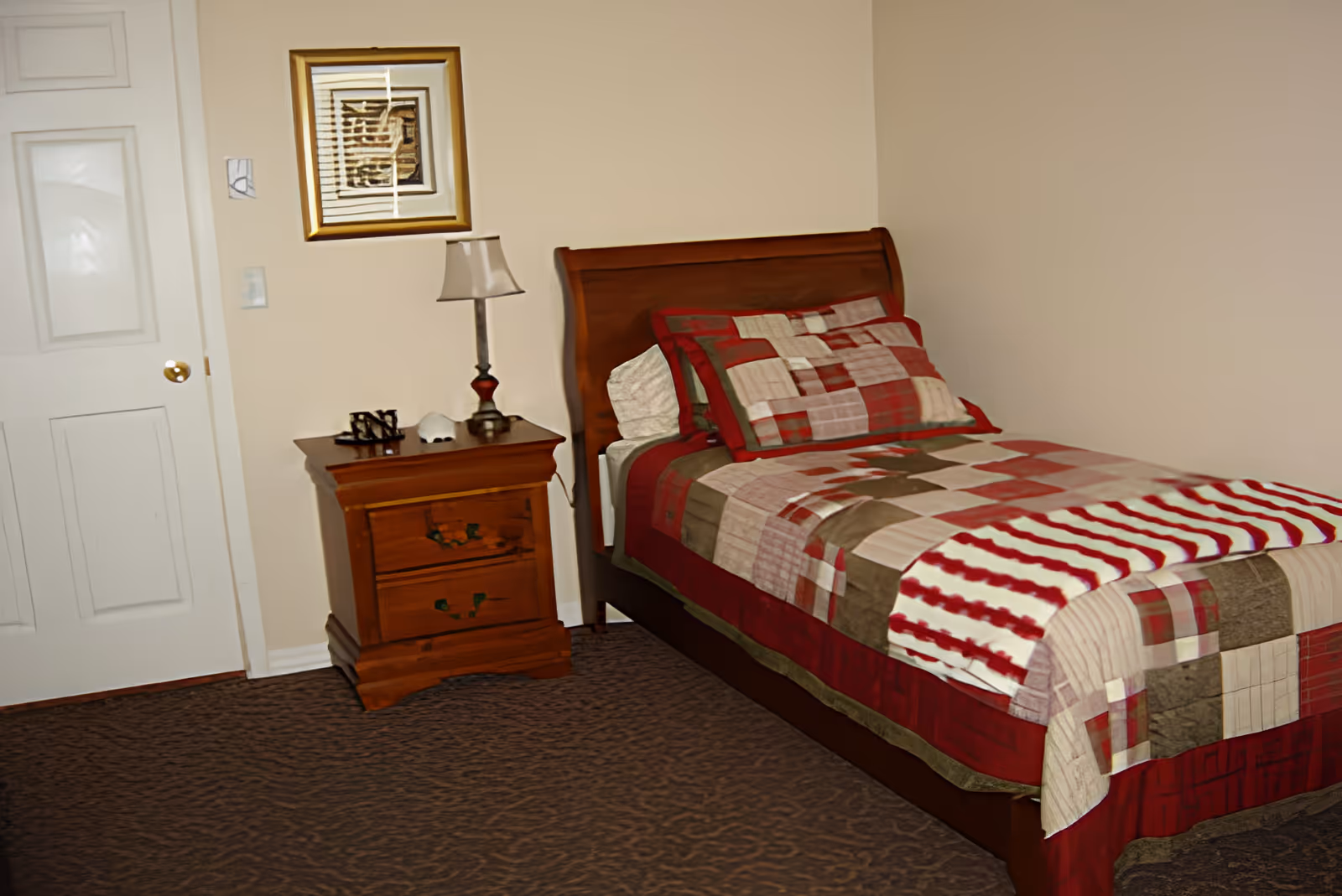 A bedroom with a single wooden bed covered with a red, beige, and brown patchwork quilt and matching pillow. Next to the bed is a wooden nightstand with two drawers, a small lamp, and decorative items. A framed artwork hangs on the beige wall above the nightstand. The room has a closed white door and carpeted floor.