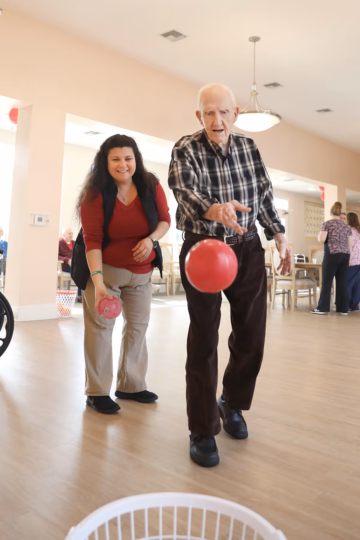 An elderly man and a woman are playing an indoor bowling game in a bright room with wooden floors. The man is rolling a red ball towards a white basket on the floor, while the woman smiles and holds another red ball behind him. Other people are visible in the background, sitting and standing near tables and chairs.