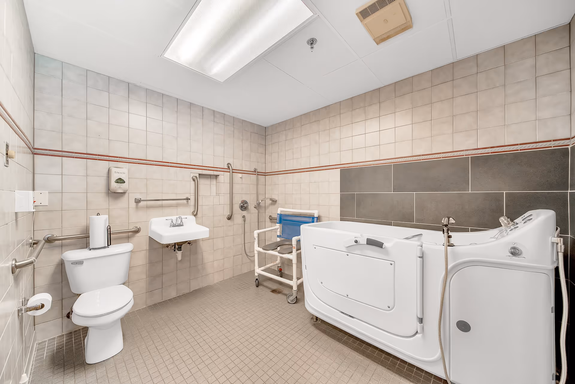 A spacious accessible bathroom with beige tiled walls and floor. The room features a white toilet with grab bars, a wall-mounted sink with grab bars, a shower area with a handheld showerhead, a blue shower chair with wheels, and a large white walk-in bathtub with controls and a handheld shower attachment. The ceiling has a large fluorescent light and a ventilation unit.