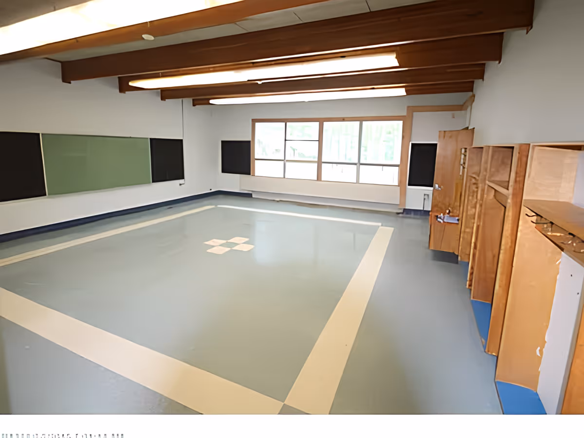 An empty indoor room with a large square area marked on the floor, possibly for activities or games. The room has wooden beams on the ceiling, large windows on one side letting in natural light, and wooden storage cabinets along one wall.