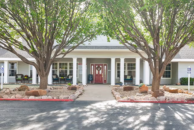 Front exterior view of a single-story building with a red door, white columns, and large windows. Two large trees with green leaves frame the entrance, and there are landscaped rock beds on either side of the walkway. Handicap parking signs are visible near the entrance.