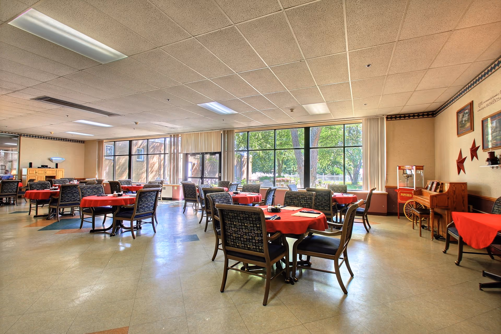 A spacious dining room with multiple round tables covered with red tablecloths and surrounded by chairs. Large windows allow natural light to fill the room, showing a view of trees outside. The room has a popcorn machine, a piano, and wall decorations including framed pictures and red star ornaments.