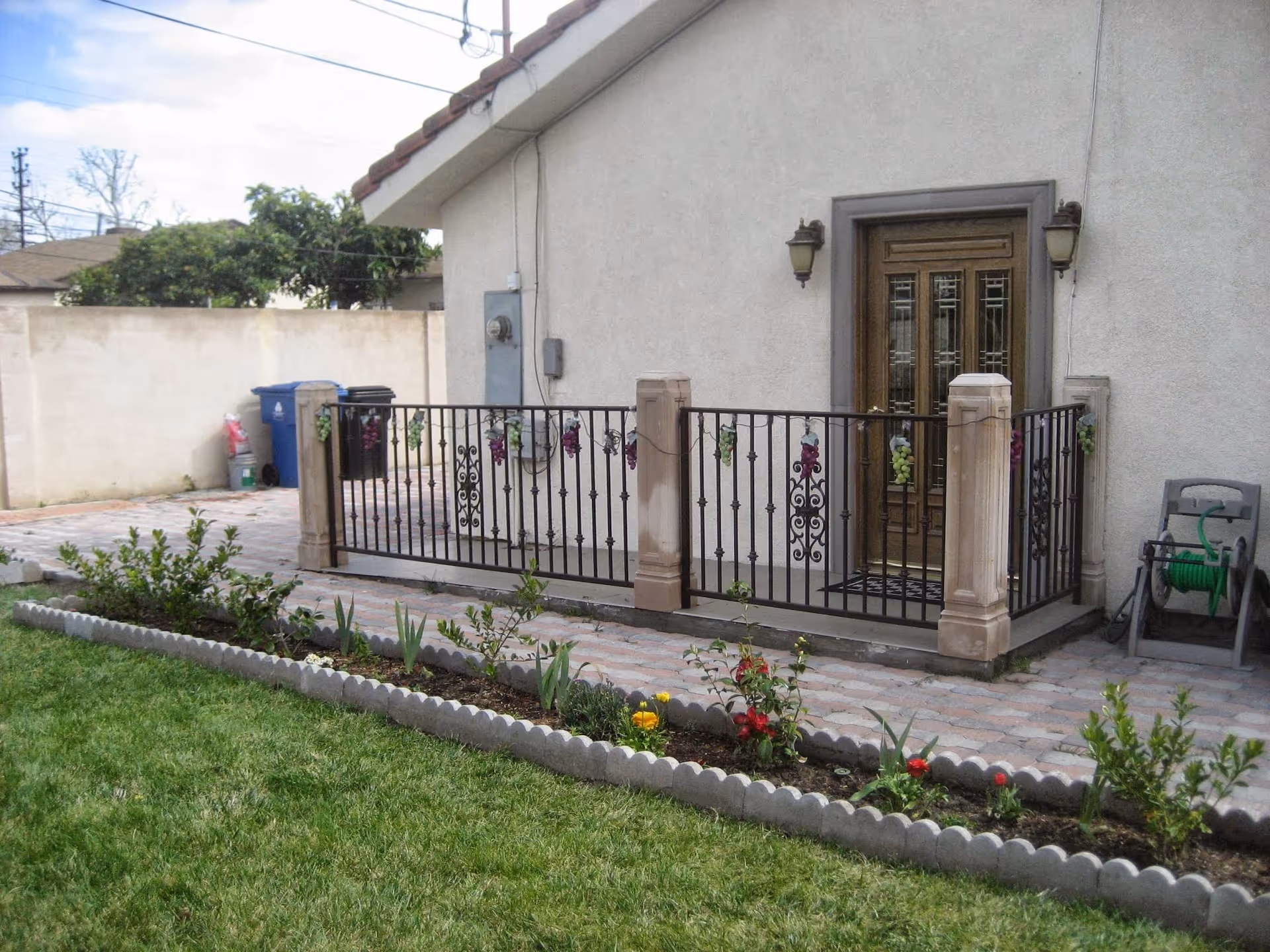 Outdoor view of a small patio area attached to a building with a decorative metal railing and a wooden door. The patio is surrounded by a paved walkway and a garden bed with various plants and flowers. A green garden hose reel is visible on the right side, and trash bins are seen in the background near a concrete wall.