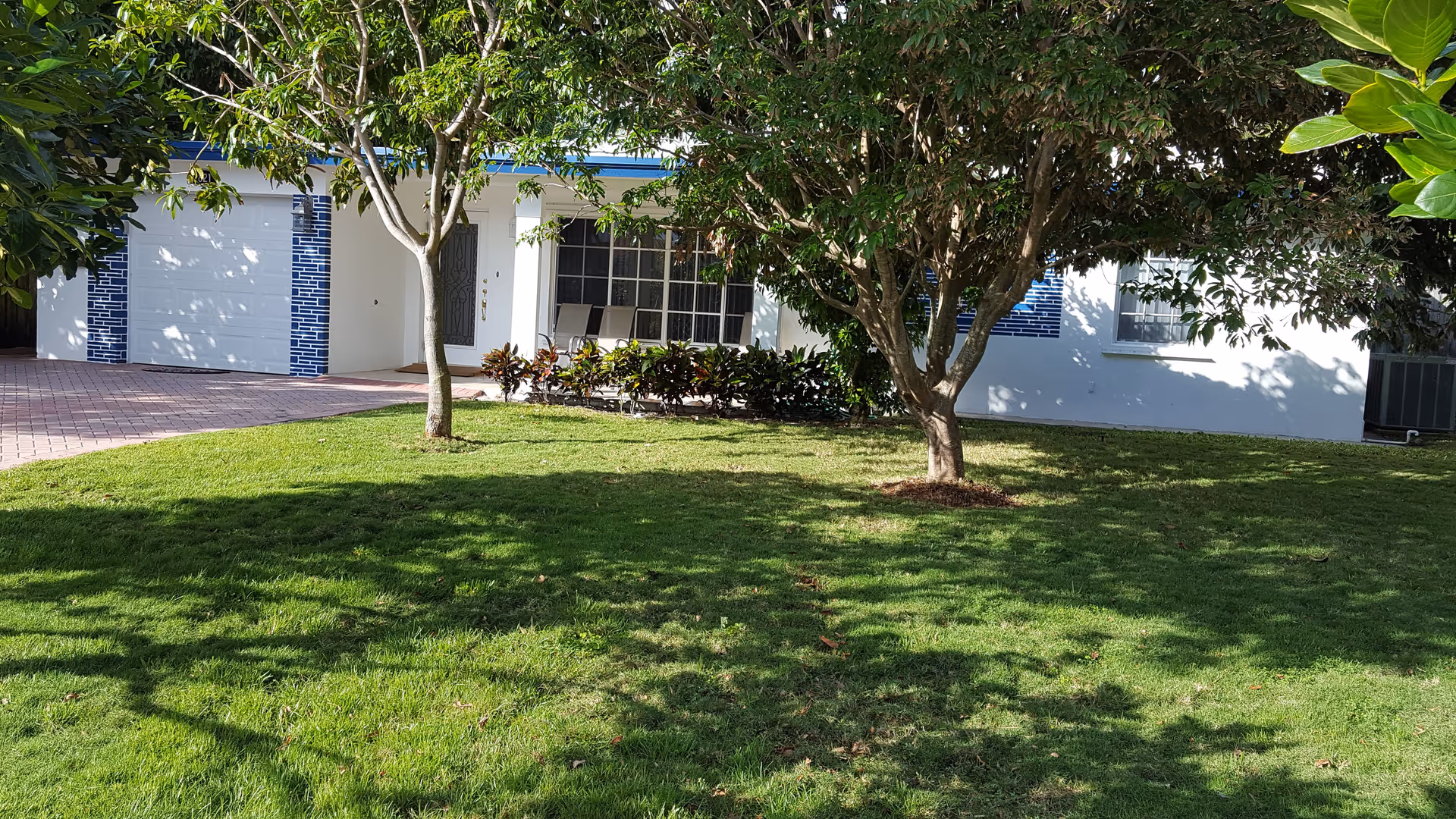 Front exterior view of a white building with blue tile accents around the garage door and windows. There is a green lawn with two trees casting shadows in the foreground. A paved driveway leads to the garage, and there are chairs visible on the porch area near the front door.