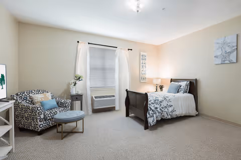 A cozy bedroom with a single bed featuring white and blue floral bedding, a bedside table with a lamp and plant, a patterned armchair with a matching ottoman, a window with white curtains and a window air conditioning unit, and light beige walls and carpet.