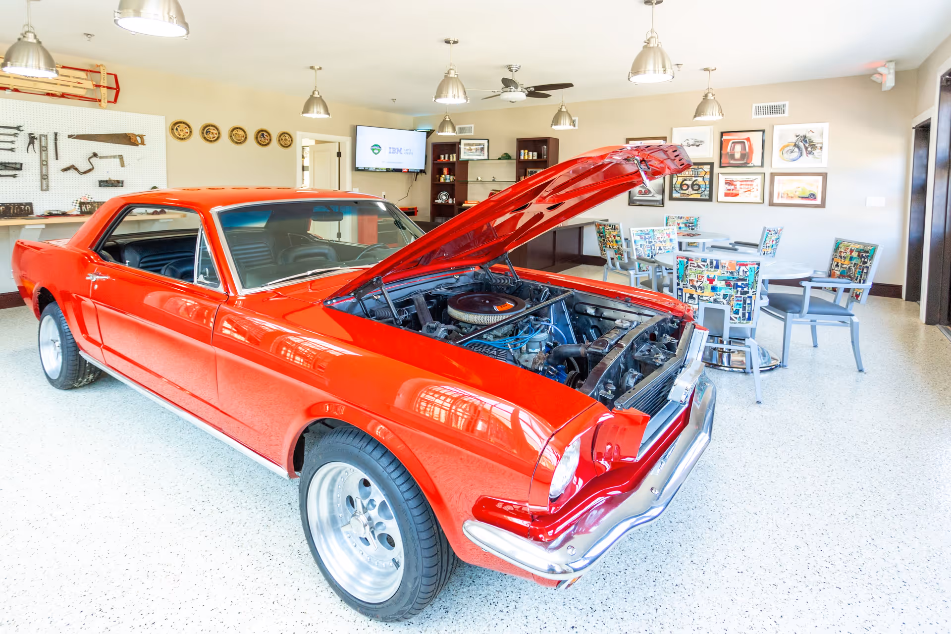 Shiny red classic car with its hood open displayed inside a bright community room with tables, chairs, and wall decor.