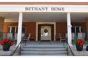 Front entrance of Bethany Home Retirement Center featuring a covered porch with white columns, two black railings on either side of the steps, two potted red flowering plants, and a glass door with sidelights.