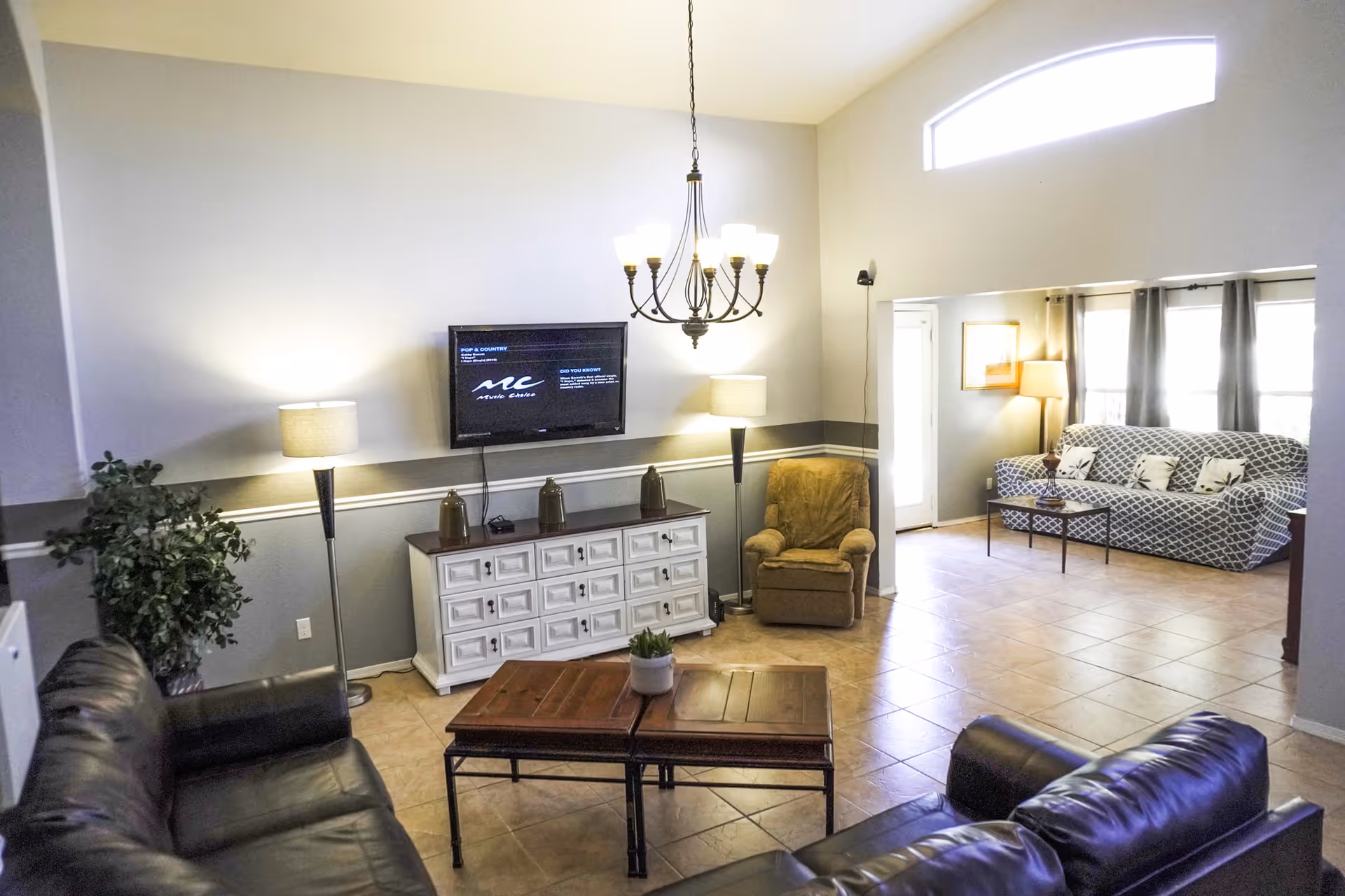 Spacious living room with leather sofas, coffee table, wall-mounted TV, chandelier, armchair, and a patterned sofa by windows.