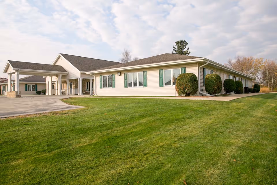 Single-story senior living facility with a covered entrance, green shutters, trimmed shrubs, and a large grassy lawn under a partly cloudy sky.