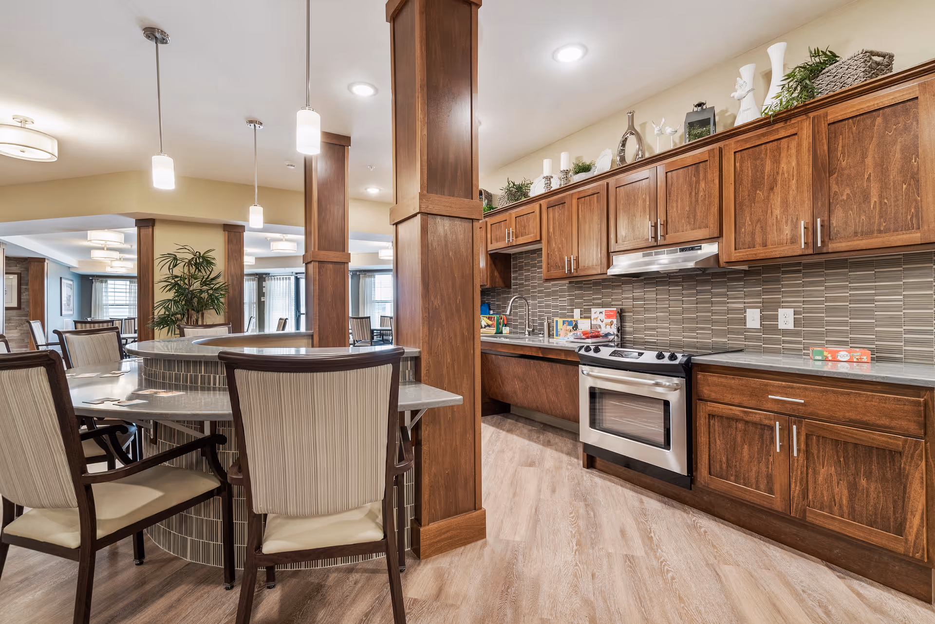 A spacious senior living facility kitchen and dining area featuring wooden cabinetry, a stainless steel oven and range hood, a tiled backsplash, and a curved dining counter with cushioned chairs. The room is well-lit with ceiling lights and pendant lights, and there are decorative plants and items on top of the cabinets.