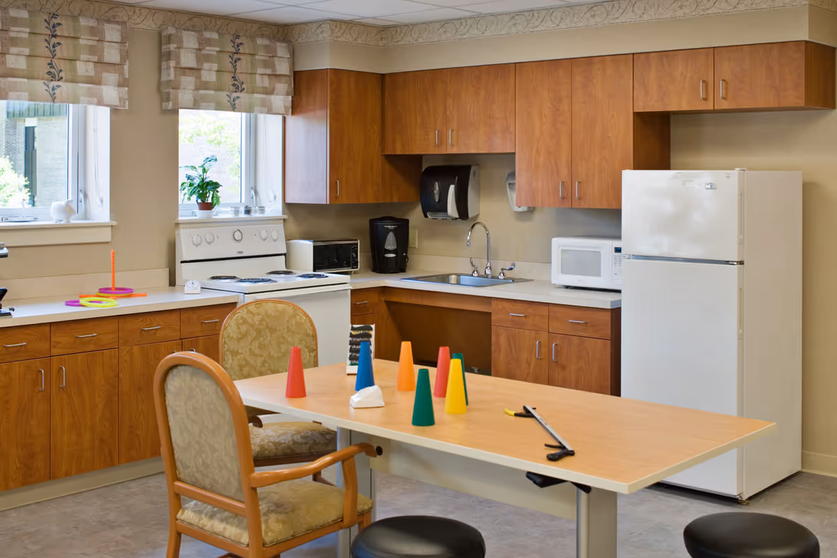 A kitchen area in a senior living facility with wooden cabinets, a white refrigerator, microwave, stove, coffee maker, and sink. There is a table in the center with colorful plastic cones and a tissue box, surrounded by chairs and stools. Two windows with patterned valances let in natural light.