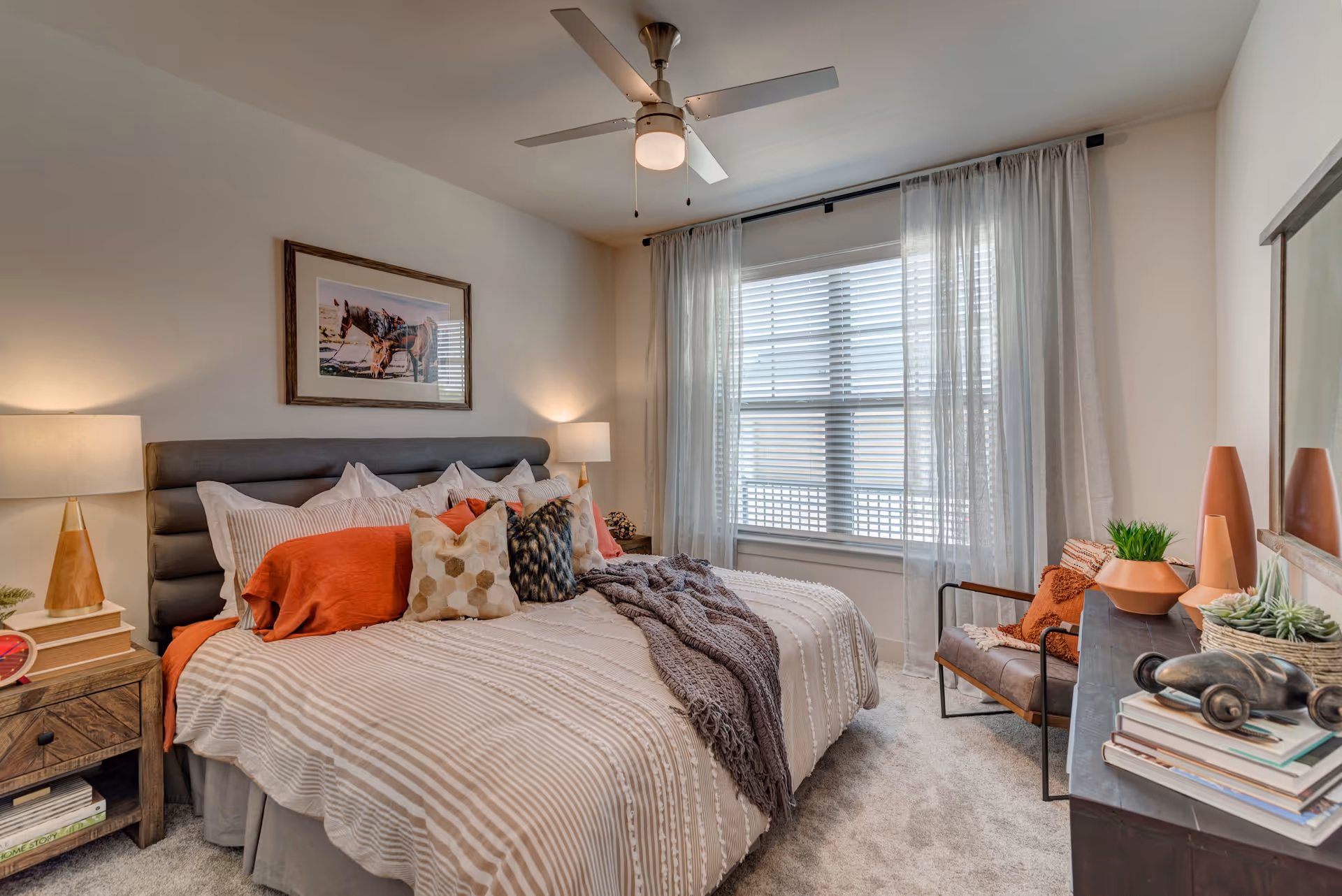 A cozy bedroom with a large bed featuring multiple pillows and a striped beige and white bedspread. There are two bedside tables with lamps, a ceiling fan with a light, a window with sheer curtains, a framed picture of a horse on the wall, and a chair with orange cushions next to a dresser decorated with plants and books.