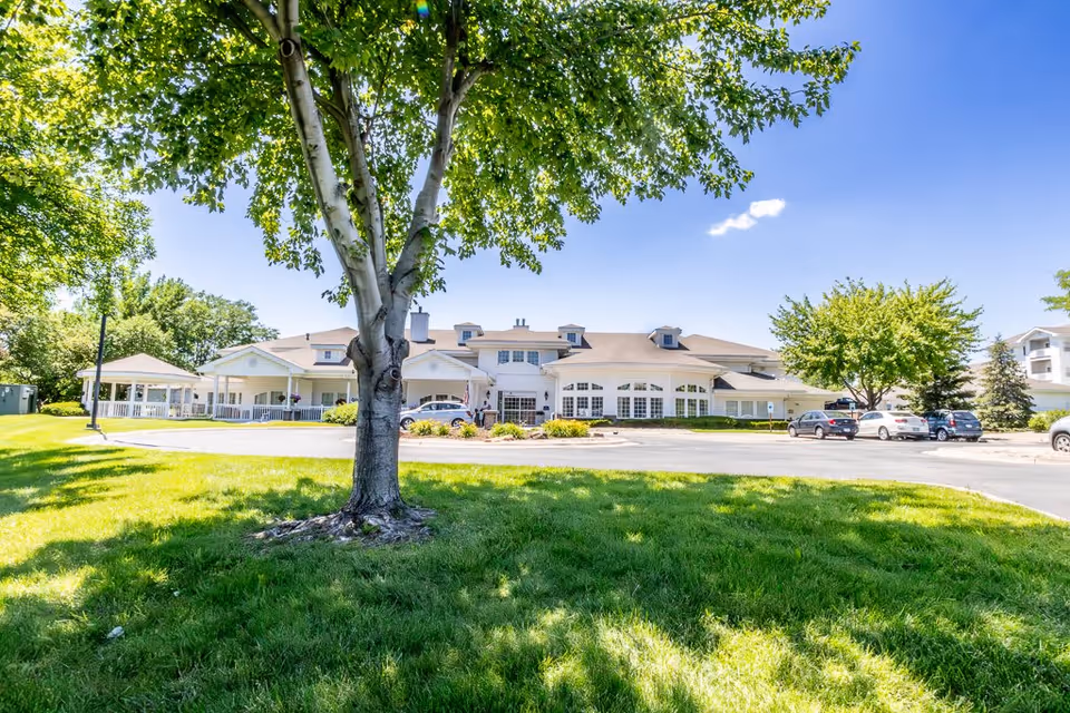 A large senior living facility building with a light-colored exterior and multiple windows, surrounded by green grass and trees under a clear blue sky. Several cars are parked in front of the building, and a large tree is prominently visible in the foreground.