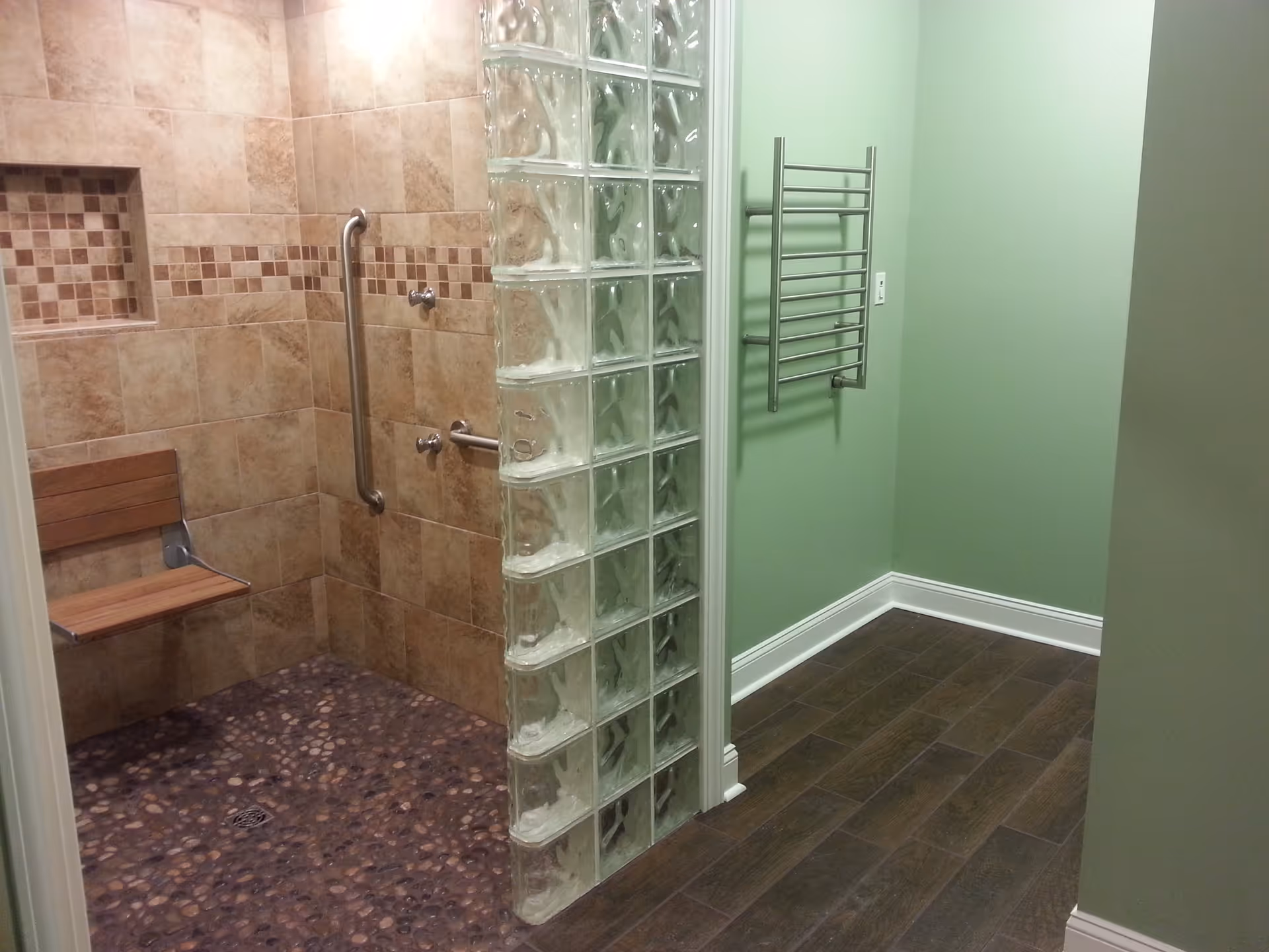 A bathroom with a walk-in shower featuring a glass block wall, tiled walls with a decorative mosaic strip, a wooden fold-down seat, and stainless steel grab bars. The adjacent area has green painted walls, a towel rack, and dark wood-look flooring.