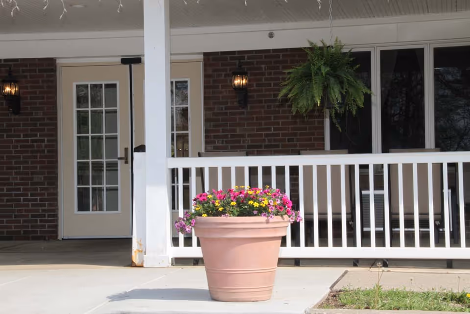 Outdoor view of a porch area with a large flower pot filled with colorful flowers in the foreground. The porch has white railings, a hanging green fern plant, two wall-mounted lantern-style lights, and a door with glass panes set in a brick wall.