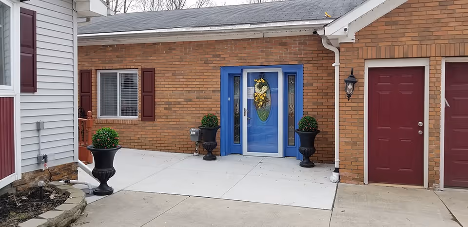 Exterior view of a senior living facility entrance with a blue door decorated with a yellow floral wreath. The building has brick walls and maroon shutters on the windows. There are two black urn planters with green plants on either side of the door, and a maroon door to the right with an outdoor wall lantern beside it. The ground is paved with concrete.