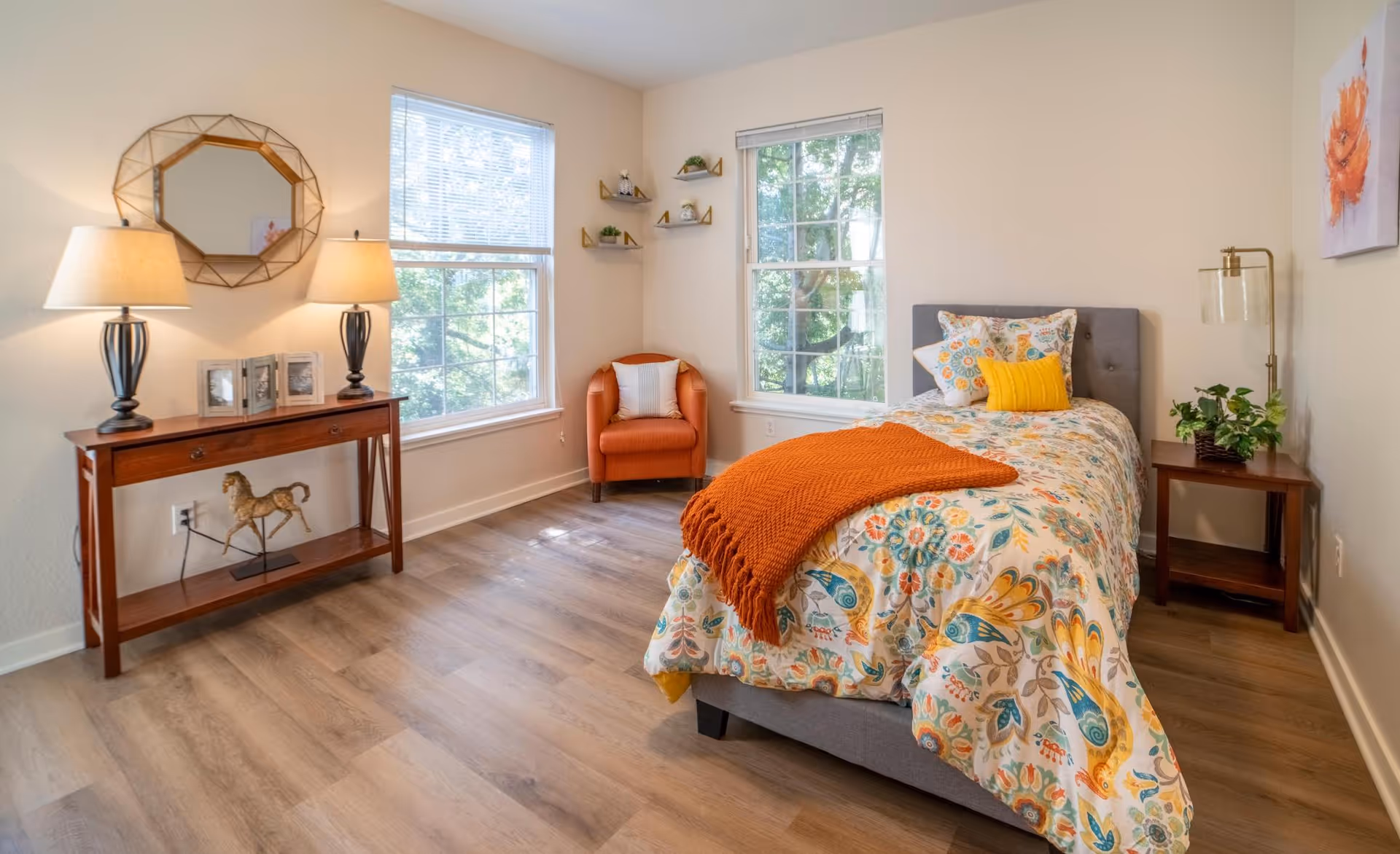 A bright bedroom with a single bed featuring a colorful floral bedspread and an orange throw blanket. There is a wooden nightstand with a lamp and a plant next to the bed. Two large windows let in natural light. An orange armchair is placed in the corner near the windows. A wooden console table with two lamps, framed photos, and a decorative horse figurine is against one wall, with a geometric mirror hanging above it. The room has light-colored walls and wood flooring.
