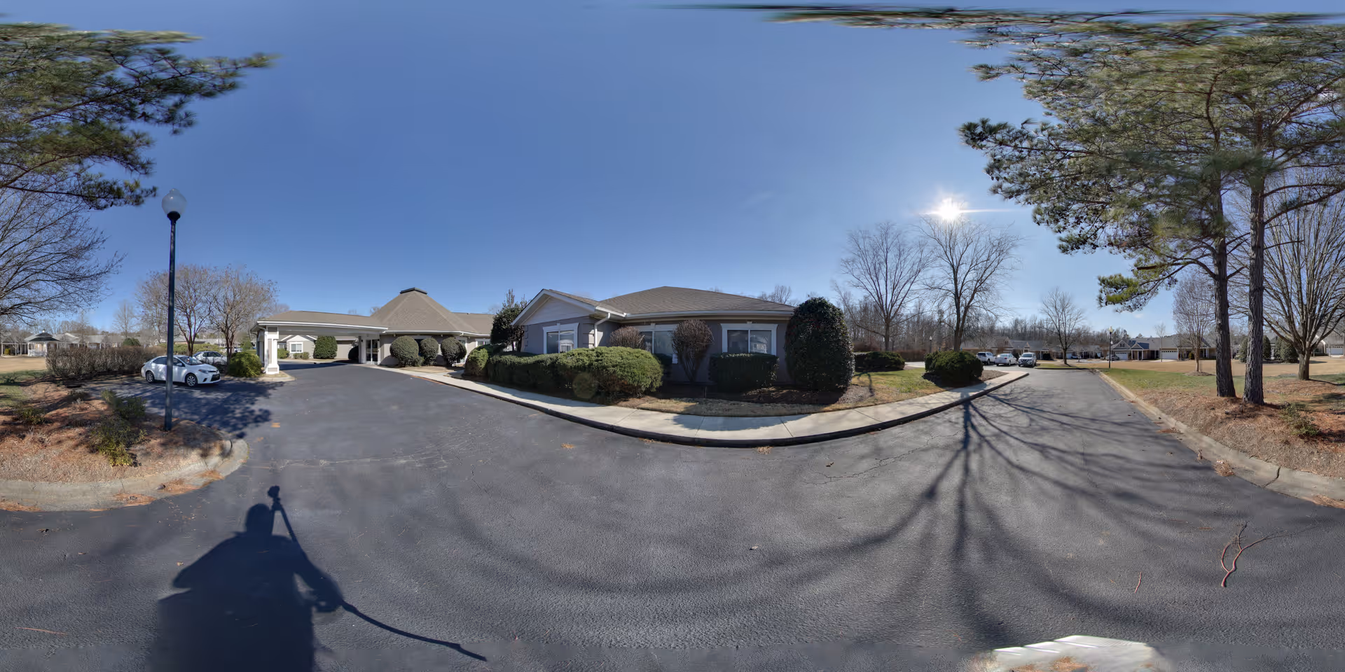 Wide panoramic view of the front exterior of a single-story senior living facility building with a driveway, landscaped bushes, trees, and a clear blue sky.