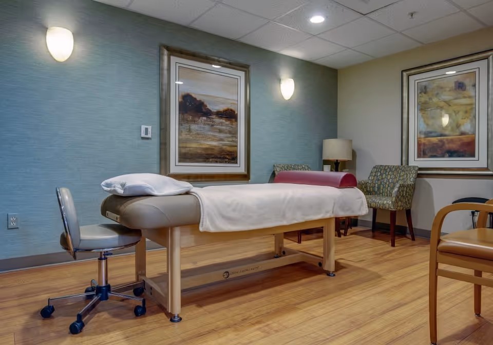A therapy or massage room with a padded treatment table covered with a white sheet and a pillow. There is a rolling chair next to the table, two patterned armchairs, a wooden chair, a floor lamp, and two framed landscape paintings on the walls. The room has wooden flooring and soft lighting from wall sconces.