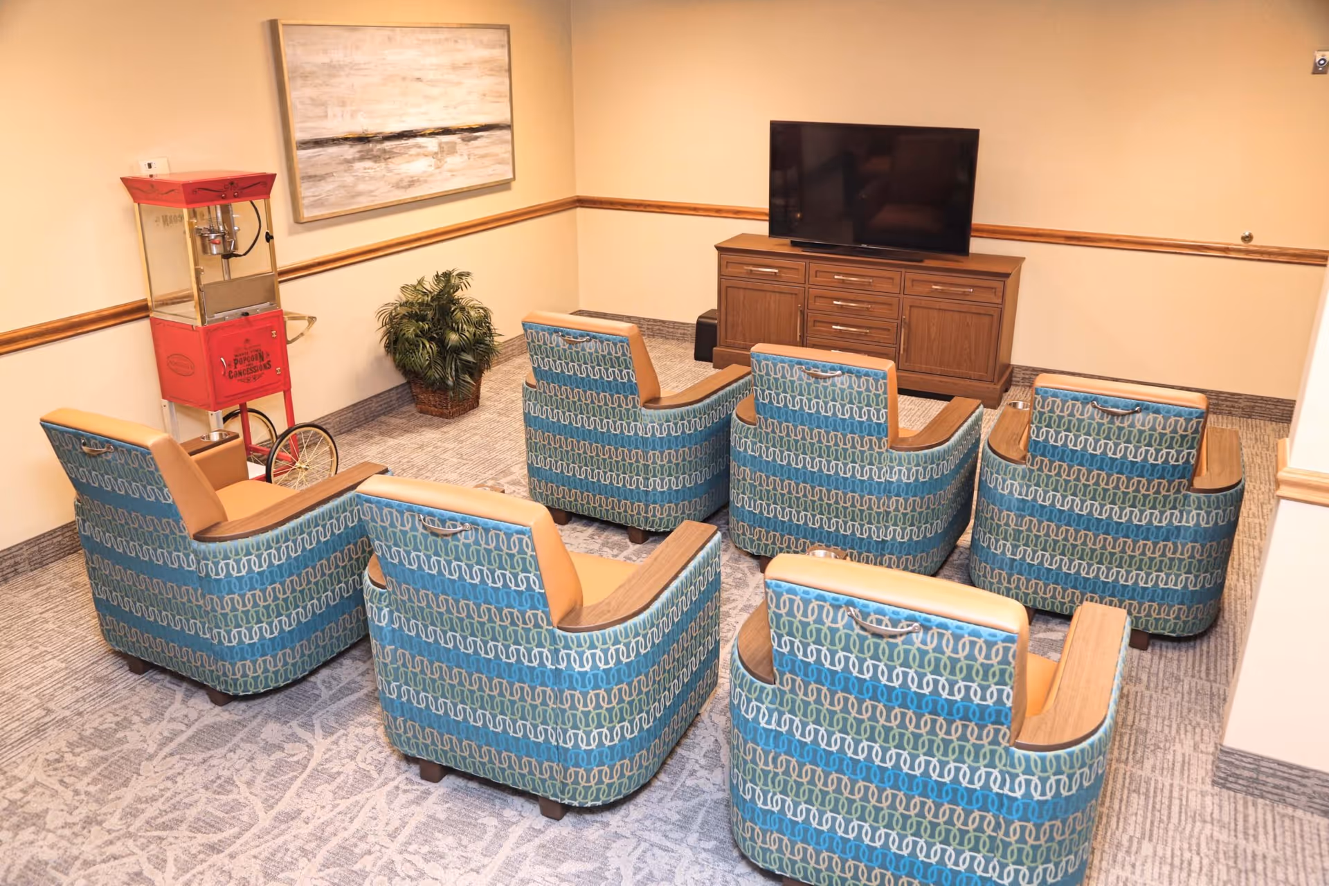 A cozy media room with six patterned armchairs arranged in two rows facing a flat-screen TV on a wooden cabinet. A red popcorn machine and a potted plant are positioned against the beige walls, which are decorated with a large abstract painting. The room has carpeted flooring and wood trim along the walls.