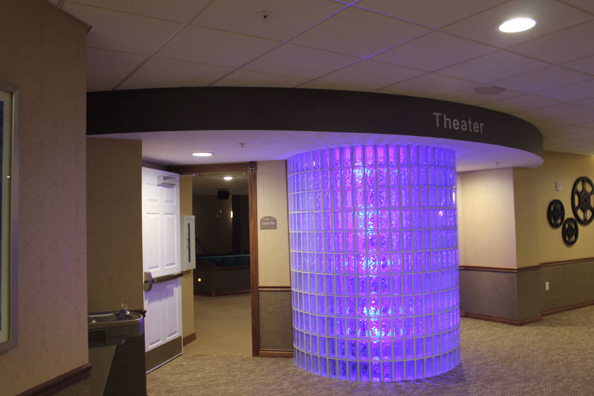 Interior hallway of a senior living facility with a curved glass block wall illuminated with purple lighting. A sign above the entrance reads 'Theater' and the door to the theater is open, showing a glimpse of theater seating inside. The hallway has beige walls with wood trim and decorative film reel wall art.