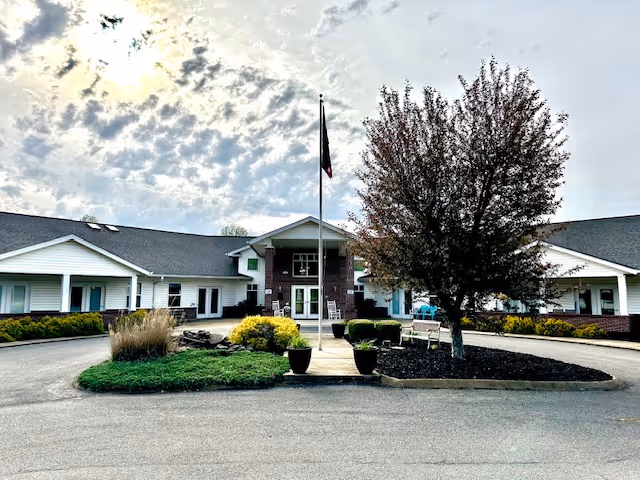 Front exterior view of a single-story assisted living facility building with a central entrance, a flagpole with a flag, landscaped greenery, and a tree in front under a partly cloudy sky.
