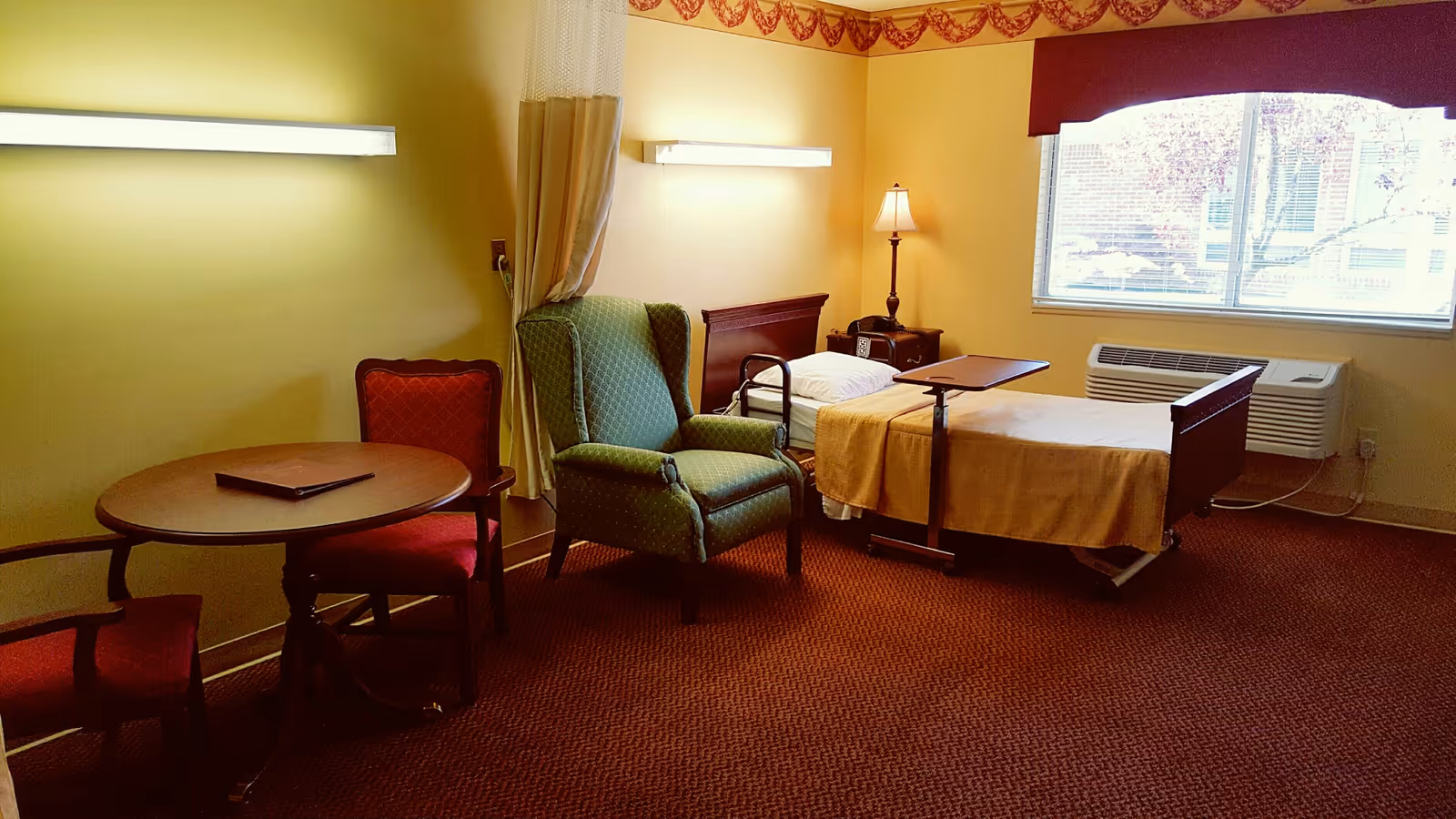 A room with a hospital-style bed covered with a beige blanket, a green upholstered armchair, a small wooden table with two red cushioned chairs, a bedside table with a lamp, and a large window with blinds letting in natural light.