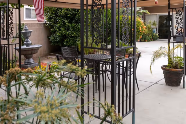 Courtyard patio with a metal gazebo, round table and chairs, potted plants and a decorative fountain by a building entrance.