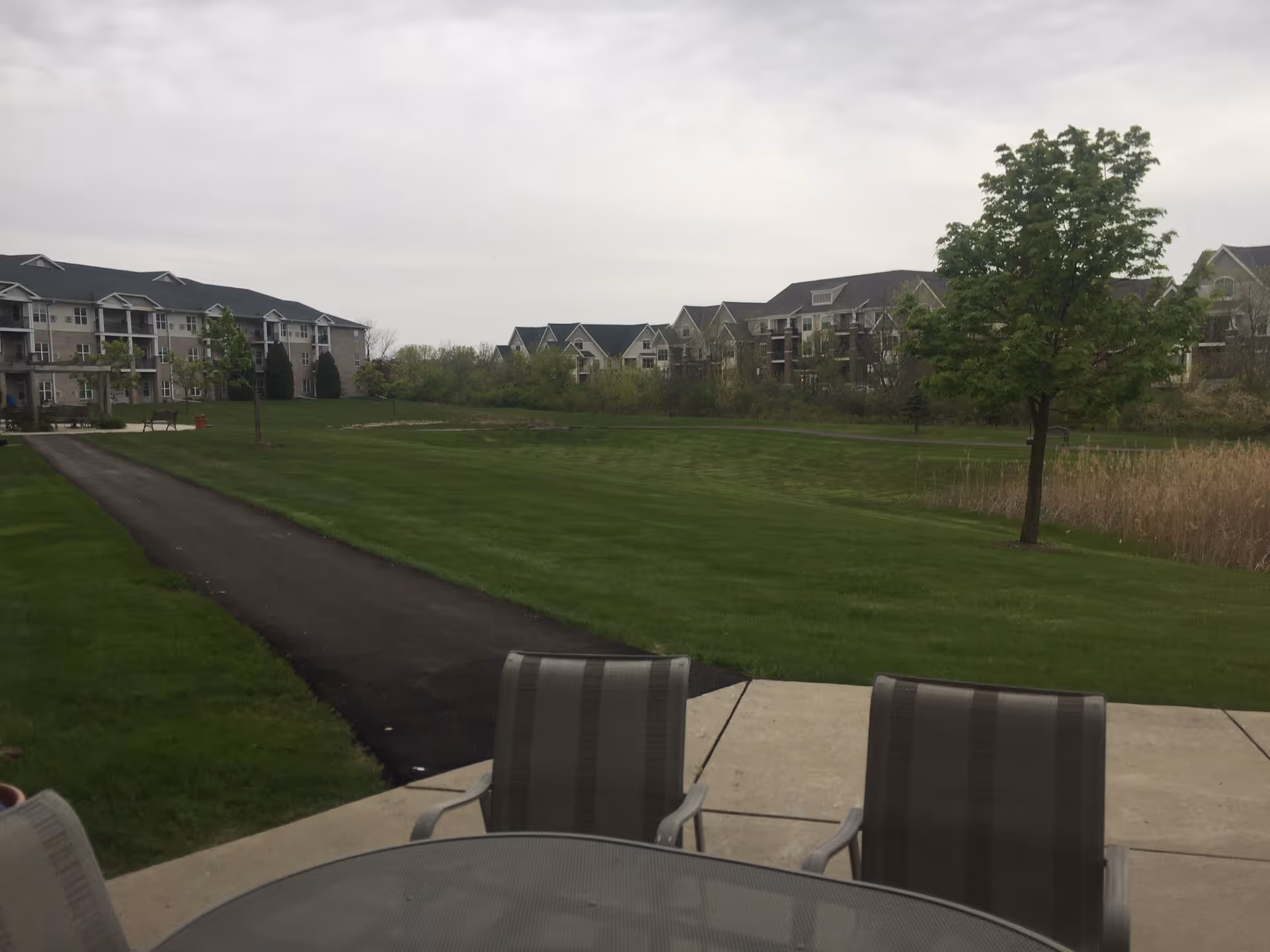 Patio table and chairs overlooking a grassy lawn with a paved path and multi-story senior living buildings under a cloudy sky.