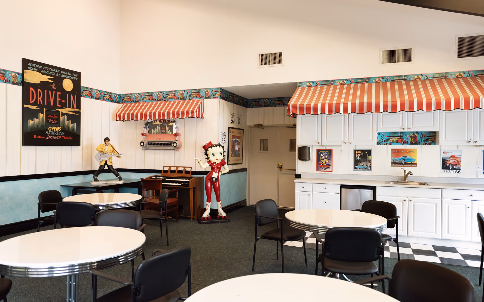 Retro-themed communal dining area with round tables and chairs, a small kitchenette with striped awnings, and vintage decor including a Betty Boop statue and drive-in poster.