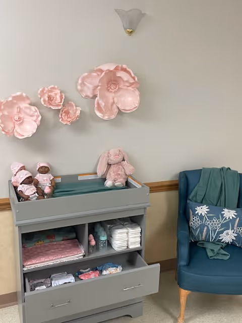 A nursery changing station with baby dolls, a pink stuffed bunny, diapers, baby clothes, and bottles. Above the station are three large pink decorative flowers mounted on the wall. To the right is a blue armchair with a floral pillow and a green blanket draped over the back.