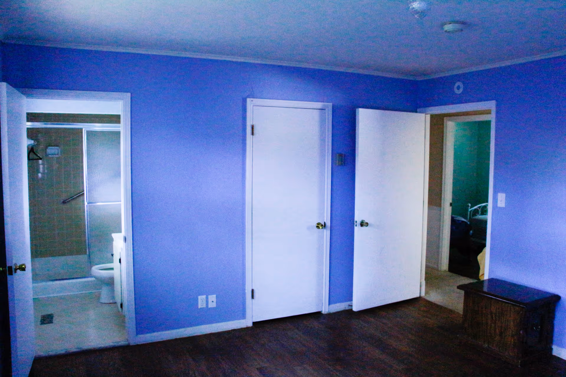 Empty purple-painted bedroom with hardwood floor, three doors (one open to a bathroom showing a toilet and shower) and a small wooden chest.