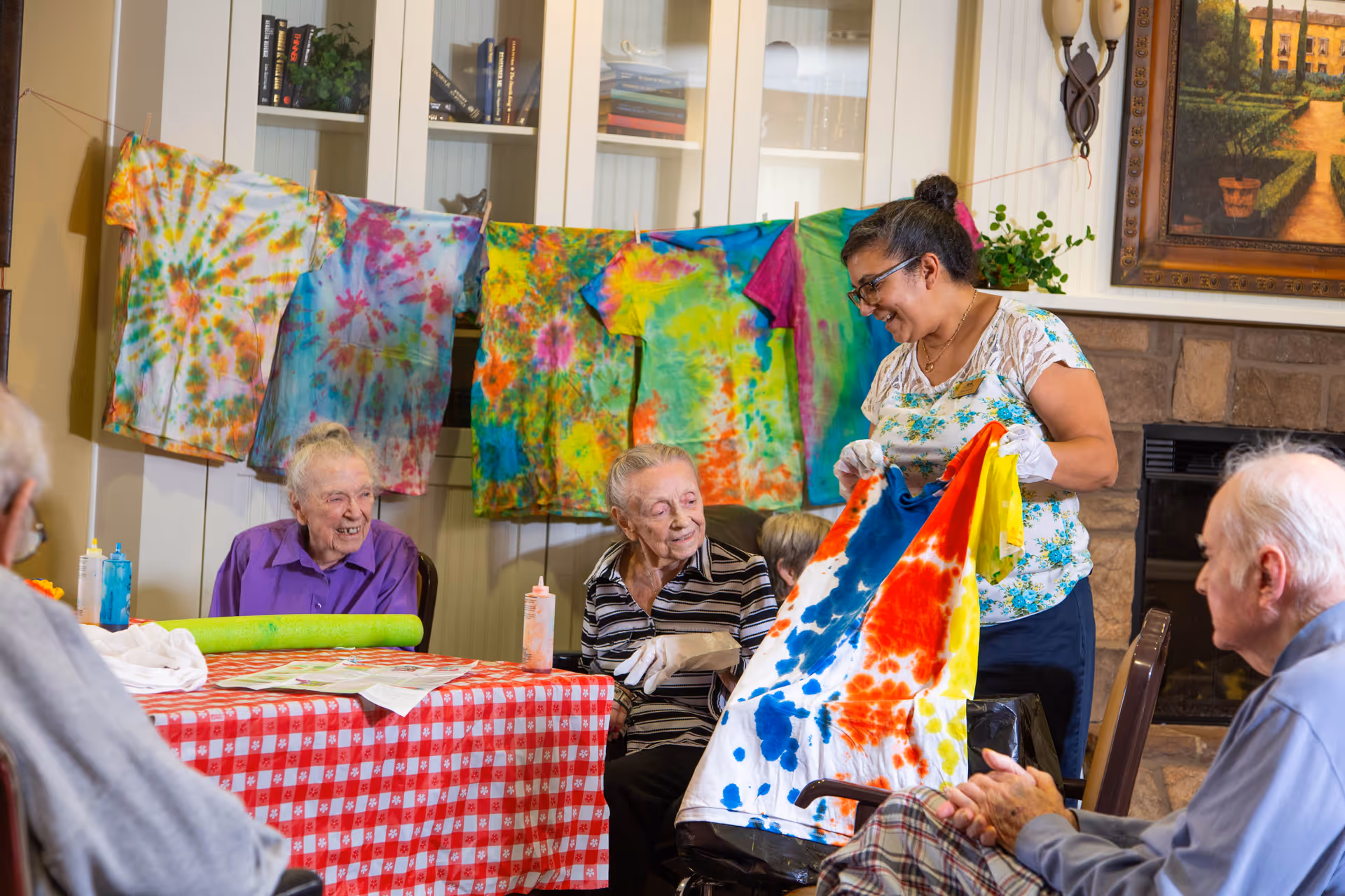 A group of elderly people sitting around a table covered with a red and white checkered tablecloth, engaging in a tie-dye activity. A staff member is standing and showing a colorful tie-dyed shirt to the group. Several tie-dyed shirts are hanging on a string in the background. The setting appears to be a cozy room with a fireplace and a painting on the wall.